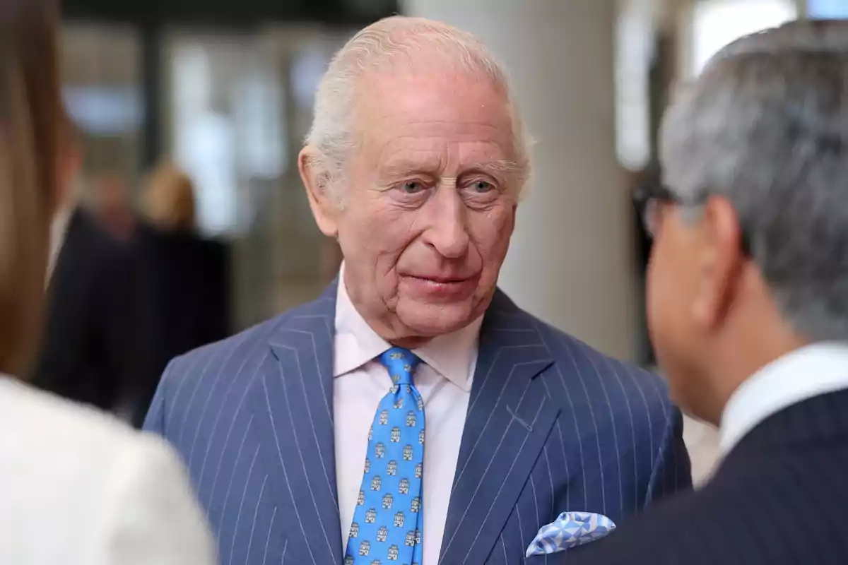 Carlos III con traje a rayas azules conversa con otra persona en un entorno interior. Carlos III con traje a rayas azules conversa con otra persona en un entorno interior.