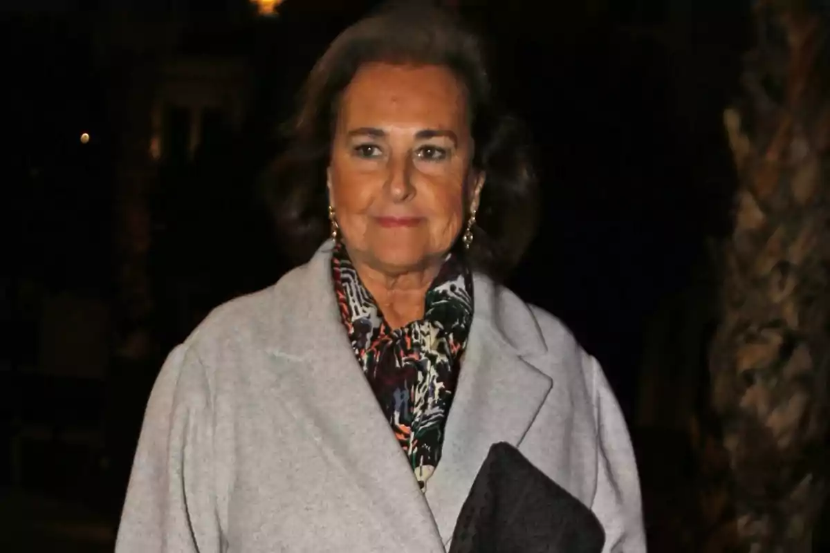 Carmen Tello, vestida con un abrigo gris y una bufanda estampada, sostiene un bolso negro mientras sonríe ligeramente en un entorno oscuro. Carmen Tello, vestida con un abrigo gris y una bufanda estampada, sostiene un bolso negro mientras sonríe ligeramente en un entorno oscuro.