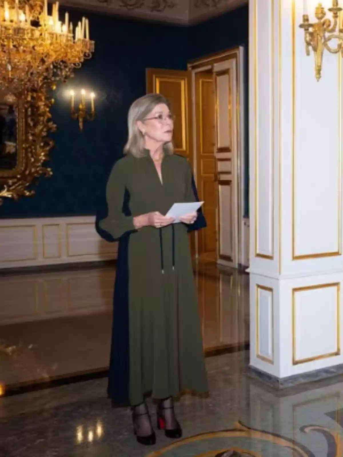 Gray-haired woman in a long dark green dress holds some papers while standing in an elegant room decorated with golden chandeliers and white walls with gold details. Gray-haired woman in a long dark green dress holds some papers while standing in an elegant room decorated with golden chandeliers and white walls with gold details.