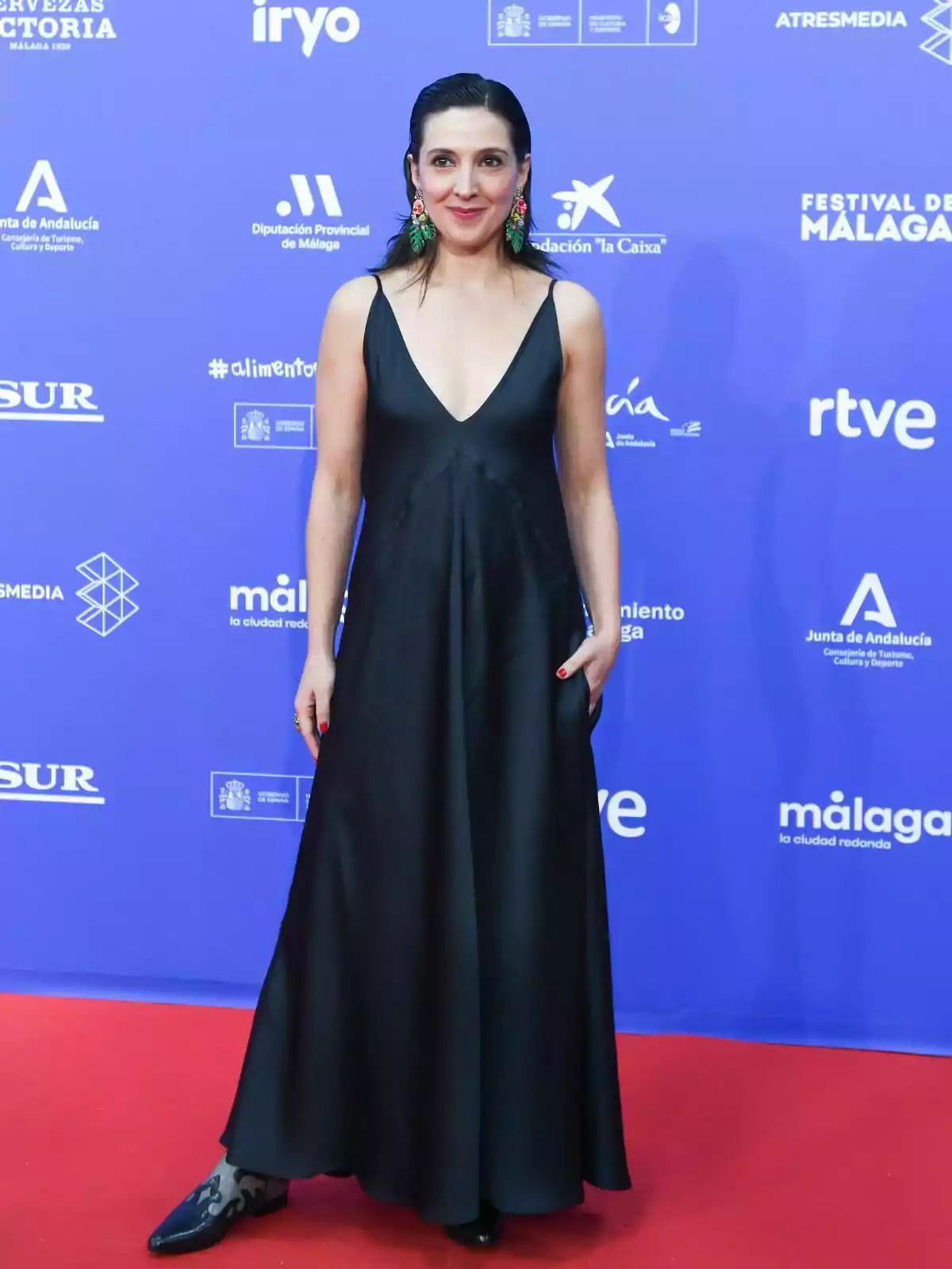 Carolina Lapausa con un vestido negro largo y aretes coloridos posa en una alfombra roja frente a un fondo azul con varios logotipos y textos.