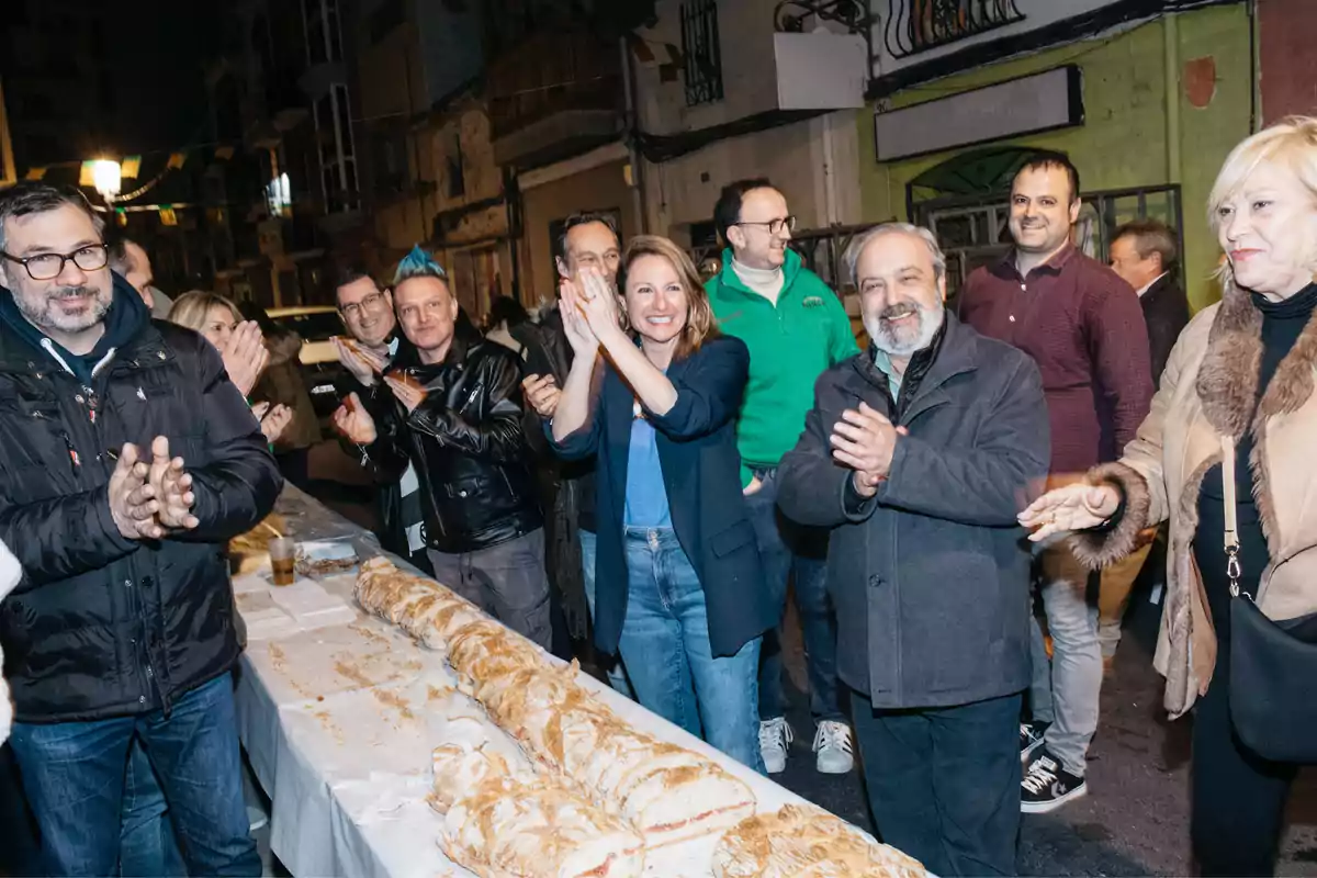 A group of people claps and smiles around a table with bread on a street lit up at night.