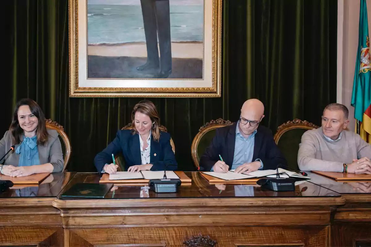 Four people sitting at a wooden table, two of them signing documents, with a large painting on the wall behind them and a flag to the right.