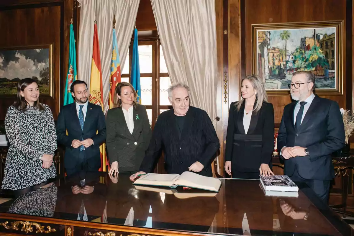 A group of people in an elegant room with flags and paintings, one of them is signing a book on a wooden table.