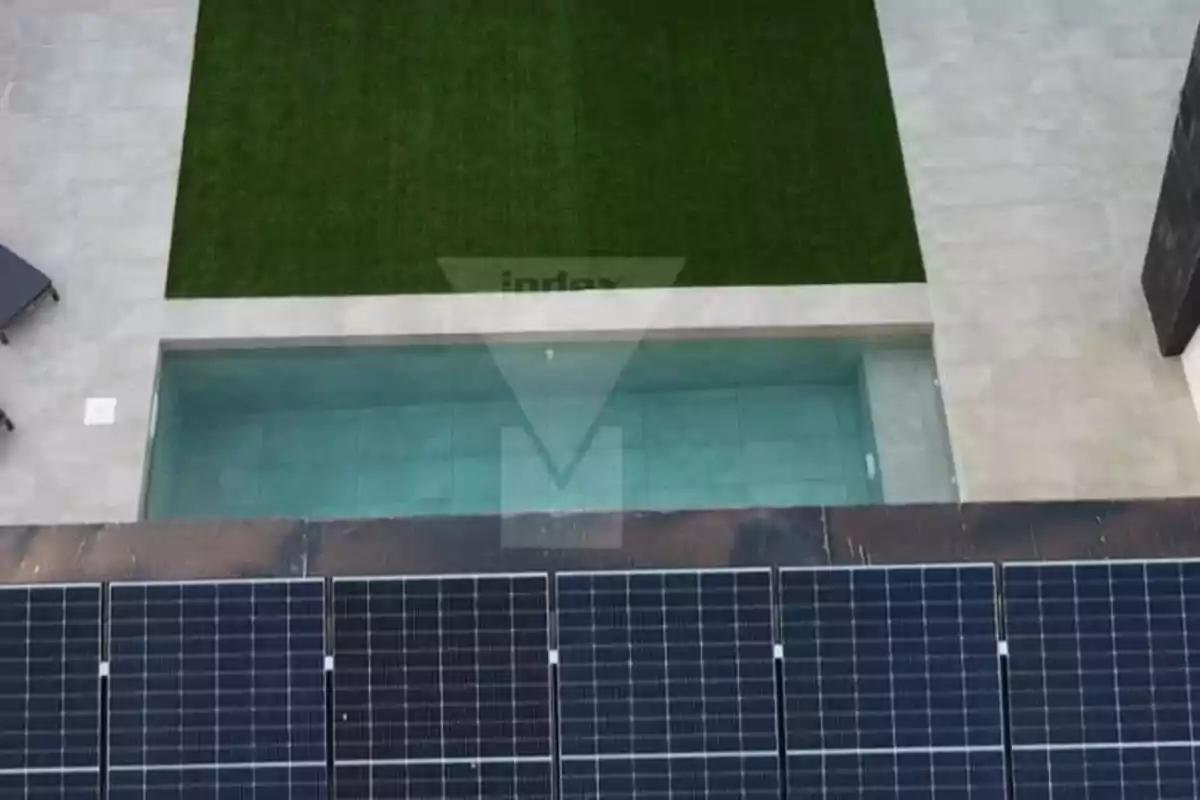 Vista aérea de un área exterior con paneles solares en primer plano, una piscina rectangular y césped artificial rodeado de pavimento claro.