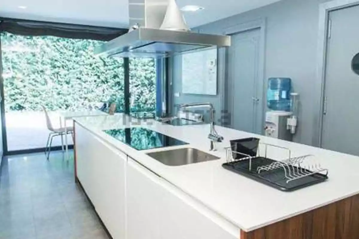 Cocina moderna con isla central blanca, fregadero, estufa de inducción, campana de acero inoxidable, escurridor de platos y dispensador de agua junto a una puerta, con ventanal al fondo que da vista a un jardín. Cocina moderna con isla central blanca, fregadero, estufa de inducción, campana de acero inoxidable, escurridor de platos y dispensador de agua junto a una puerta, con ventanal al fondo que da vista a un jardín.