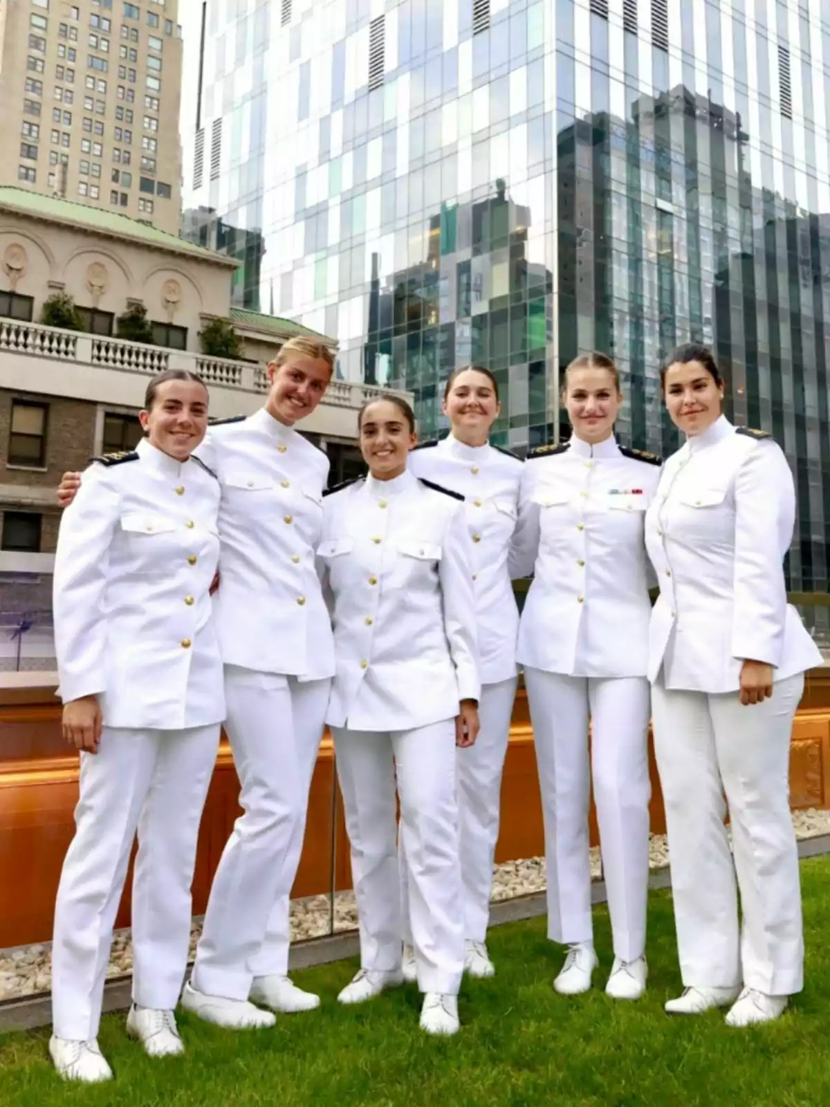 Seis mujeres vestidas con uniformes blancos de gala posan sonrientes al aire libre frente a edificios modernos y antiguos Seis mujeres vestidas con uniformes blancos de gala posan sonrientes al aire libre frente a edificios modernos y antiguos