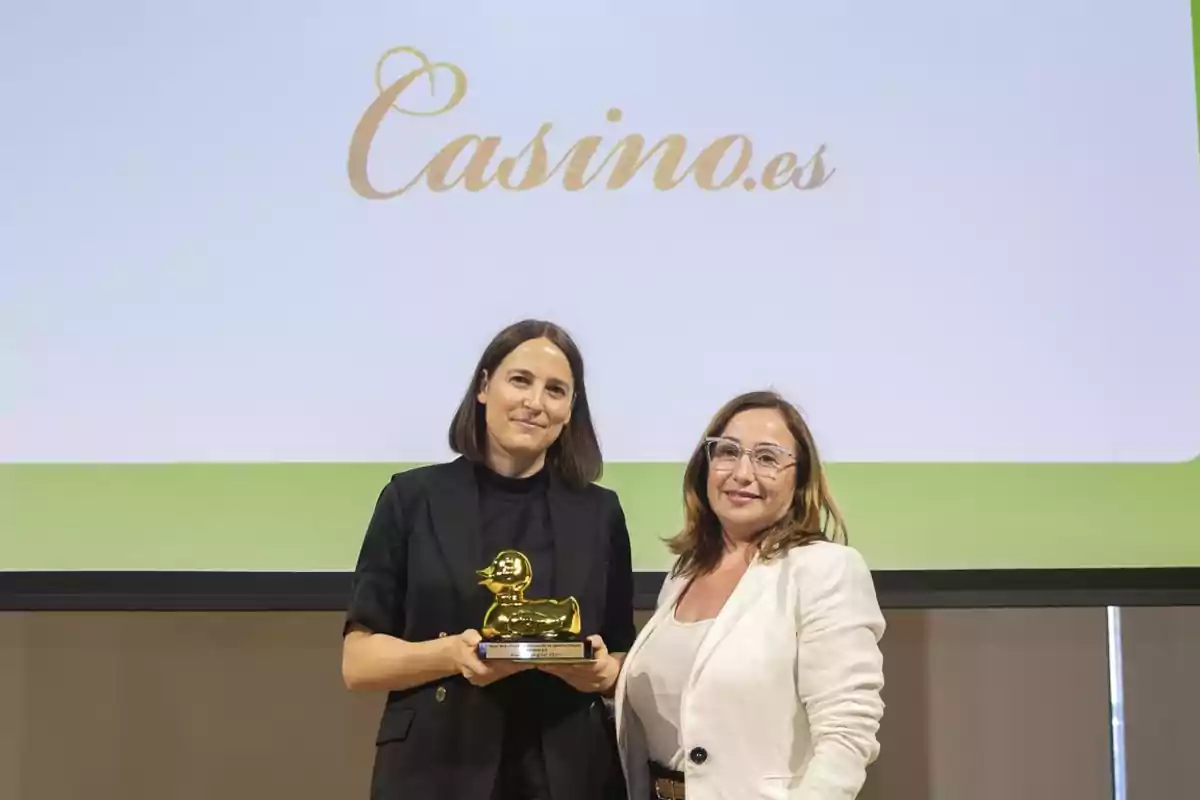 Dos mujeres posan con un trofeo dorado en forma de pato frente a una pantalla que muestra el logo de Casino.es.