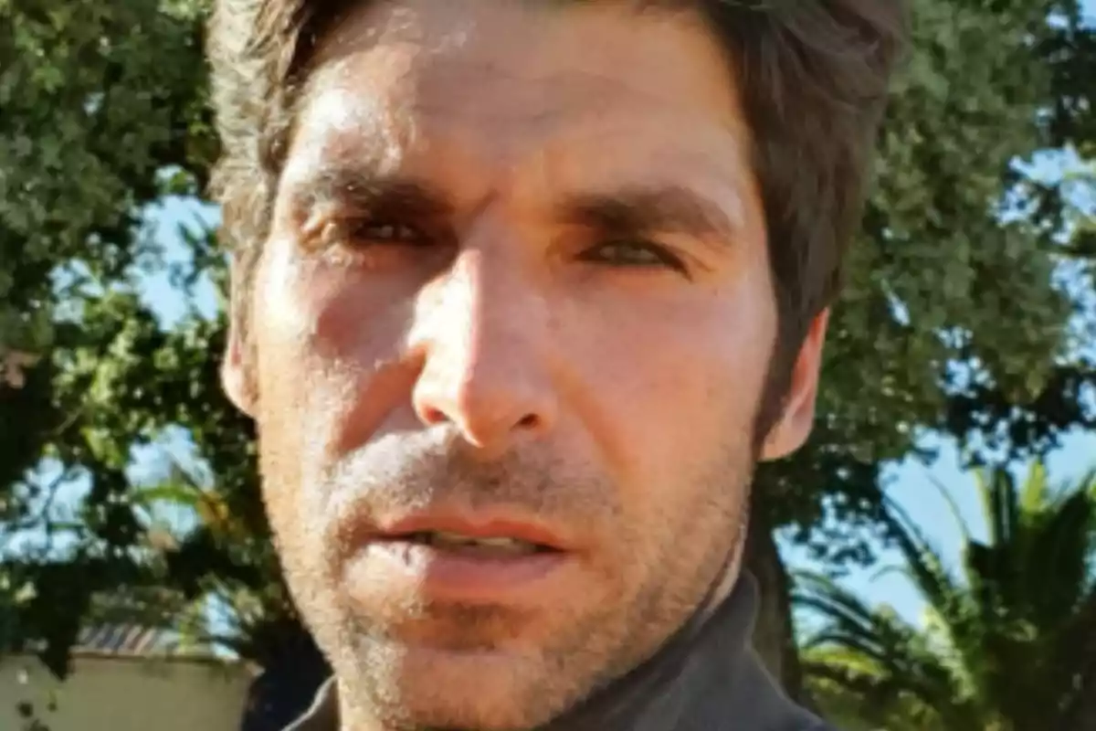 Un hombre con barba ligera y cabello oscuro en un entorno al aire libre con árboles al fondo. Un hombre con barba ligera y cabello oscuro en un entorno al aire libre con árboles al fondo.