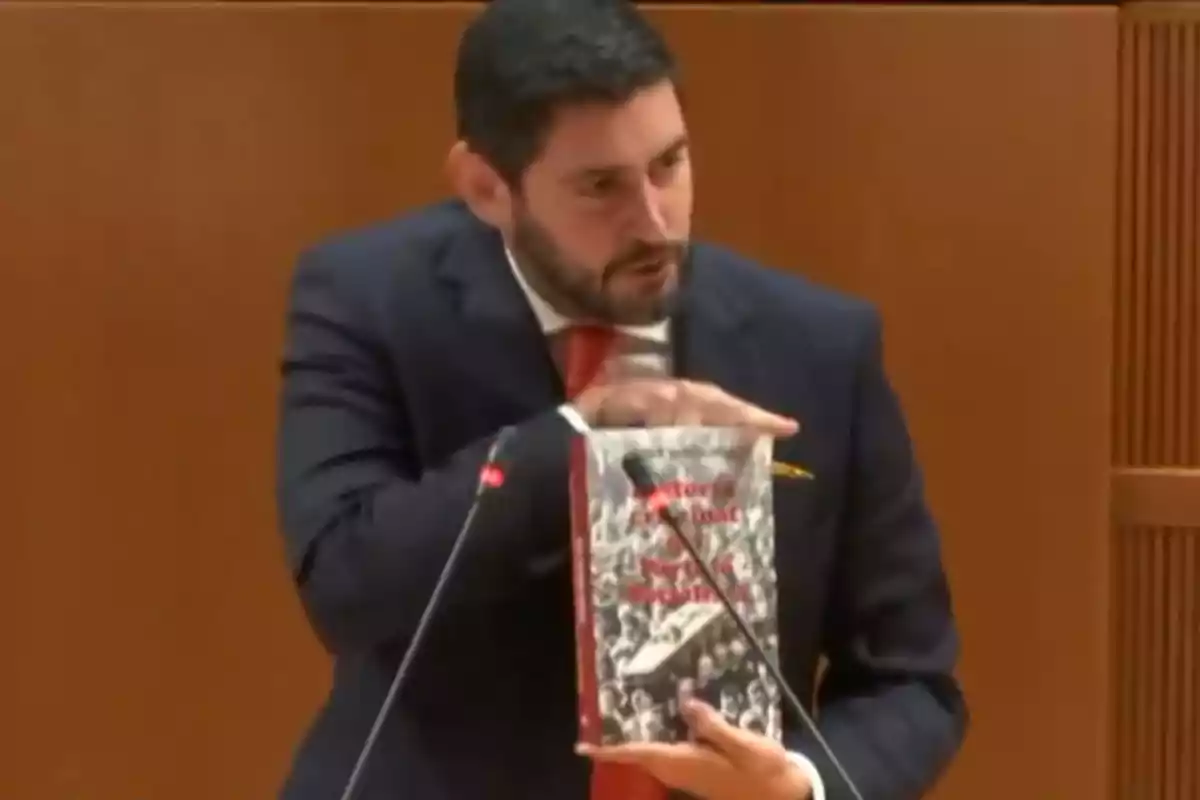 Un hombre de traje oscuro y corbata roja sostiene un libro mientras habla en un podio con micrófonos.