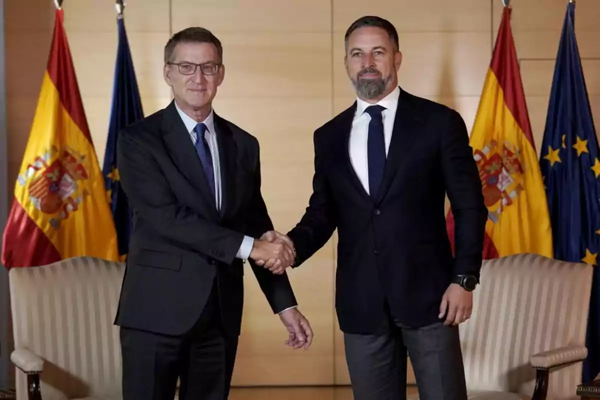 Dos hombres de traje dándose la mano con banderas de España y la Unión Europea en el fondo.