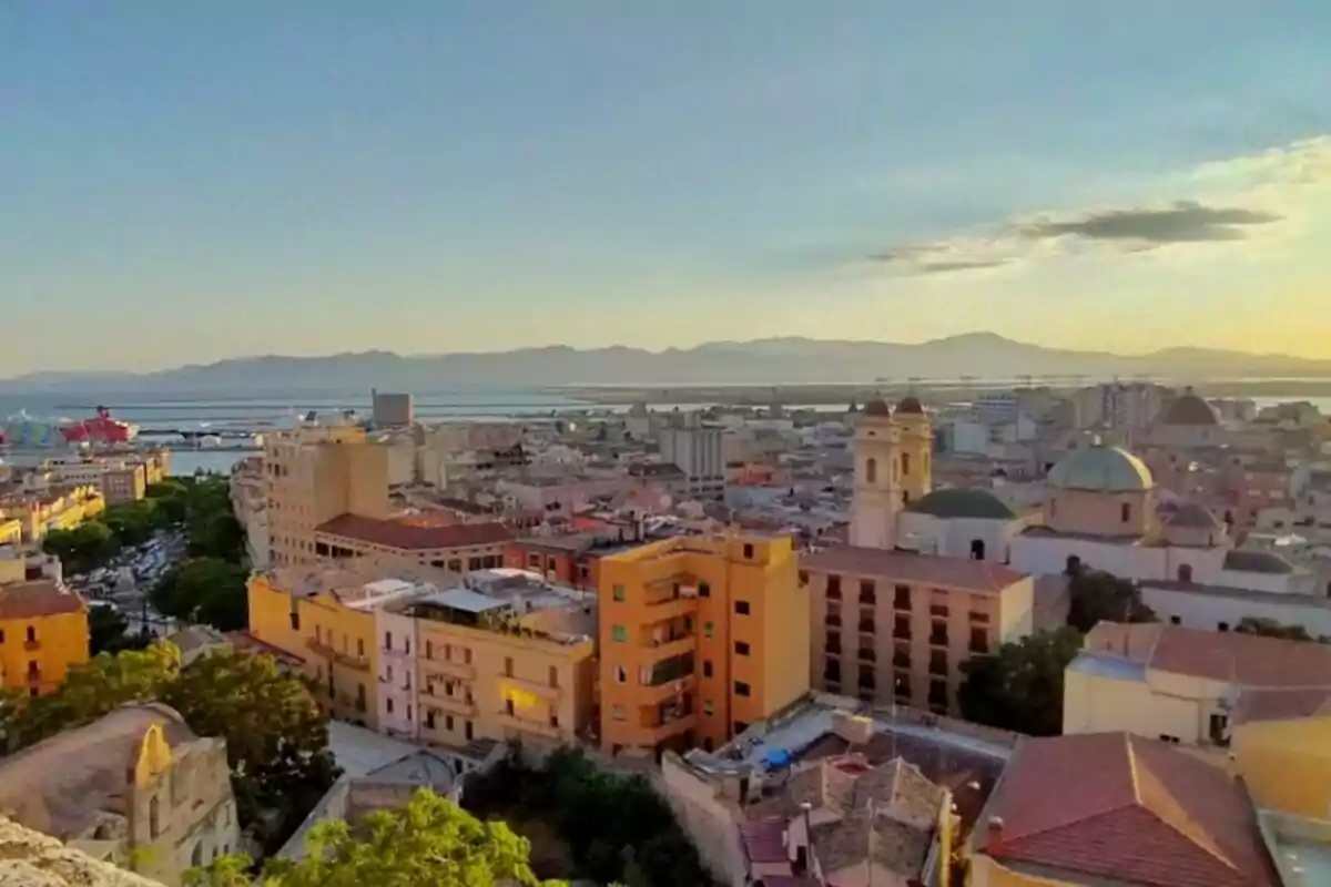 Vista panorámica de una ciudad costera al atardecer con edificios y montañas al fondo.