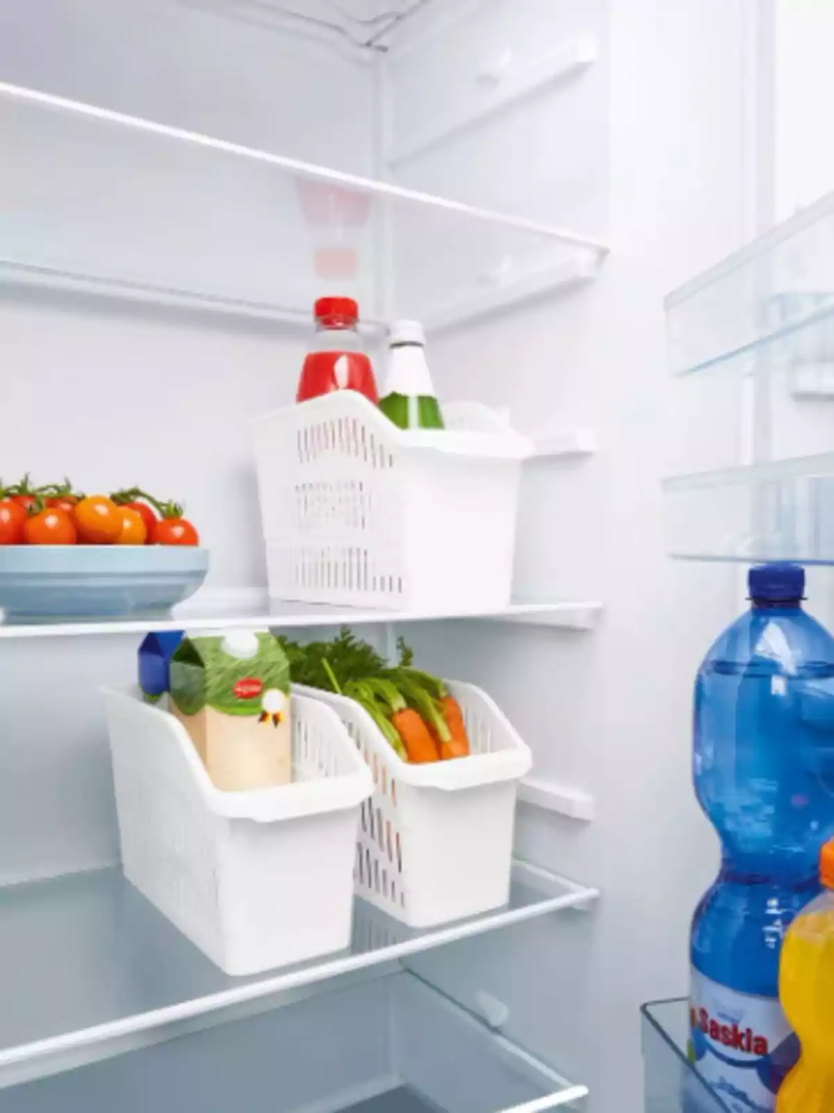 Una nevera organizada con botellas, tomates, zanahorias y un cartón de bebida en cestas blancas. Una nevera organizada con botellas, tomates, zanahorias y un cartón de bebida en cestas blancas.