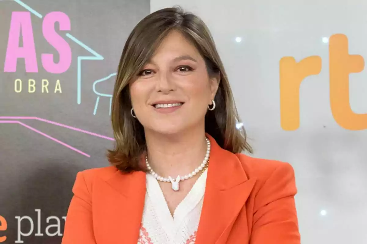 Screenshot of a smiling Chabeli Iglesias with straight brown hair, dressed in an orange blazer and a pearl necklace, poses in front of a background with logos and colorful text at the press conference for her show Los Iglesias: Hermano a la obra