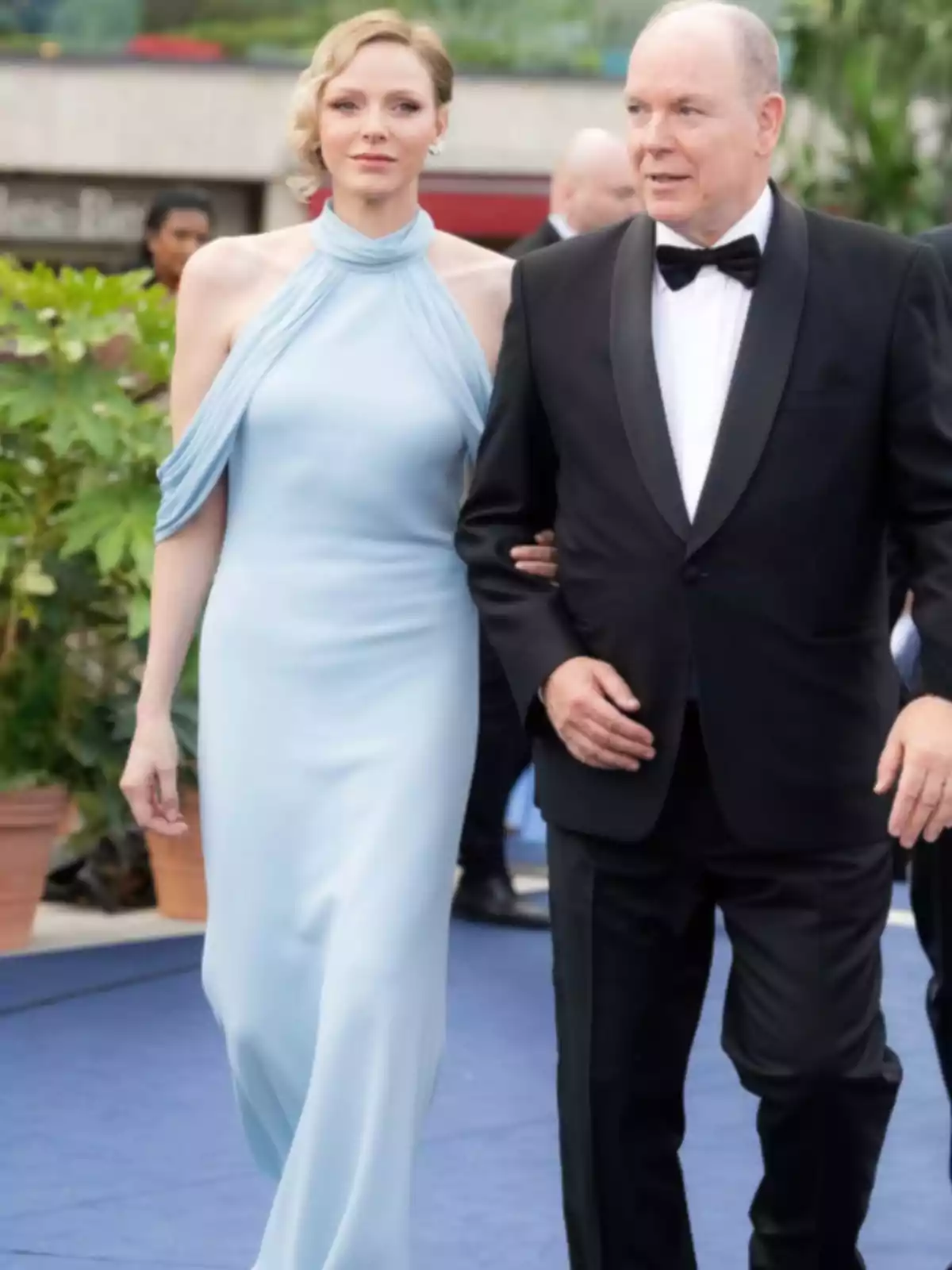 A woman in a light blue dress and a man in a black tuxedo walk together on a blue carpet at an elegant outdoor event. A woman in a light blue dress and a man in a black tuxedo walk together on a blue carpet at an elegant outdoor event.