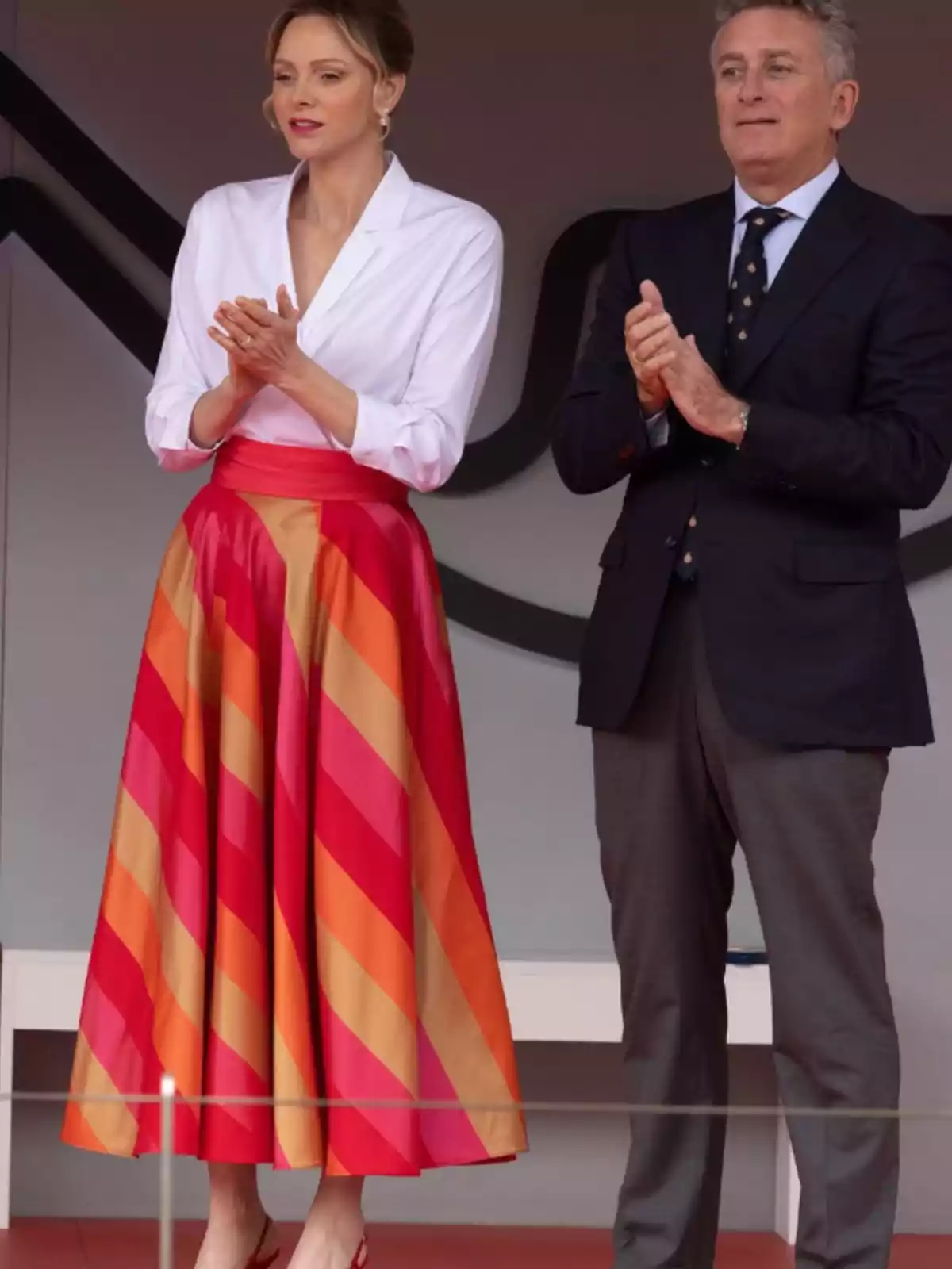 A woman in a white blouse and a red and orange striped skirt is standing next to a man in a dark suit, both applauding. A woman in a white blouse and a red and orange striped skirt is standing next to a man in a dark suit, both applauding.
