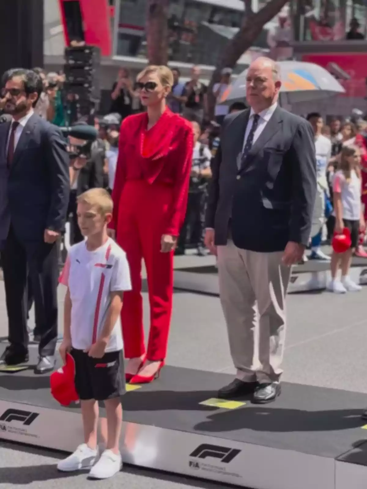 A group of people is standing on a platform during a Formula 1 event, with a child in front holding a red cap and a woman dressed in red next to a man in a suit. A group of people is standing on a platform during a Formula 1 event, with a child in front holding a red cap and a woman dressed in red next to a man in a suit.