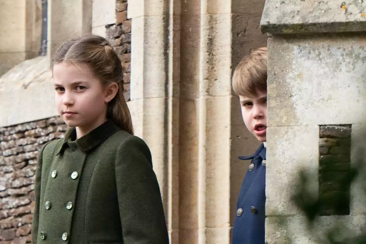 Princess Charlotte and Prince Louis in stylish coats are standing next to a stone wall. Princess Charlotte and Prince Louis in stylish coats are standing next to a stone wall.