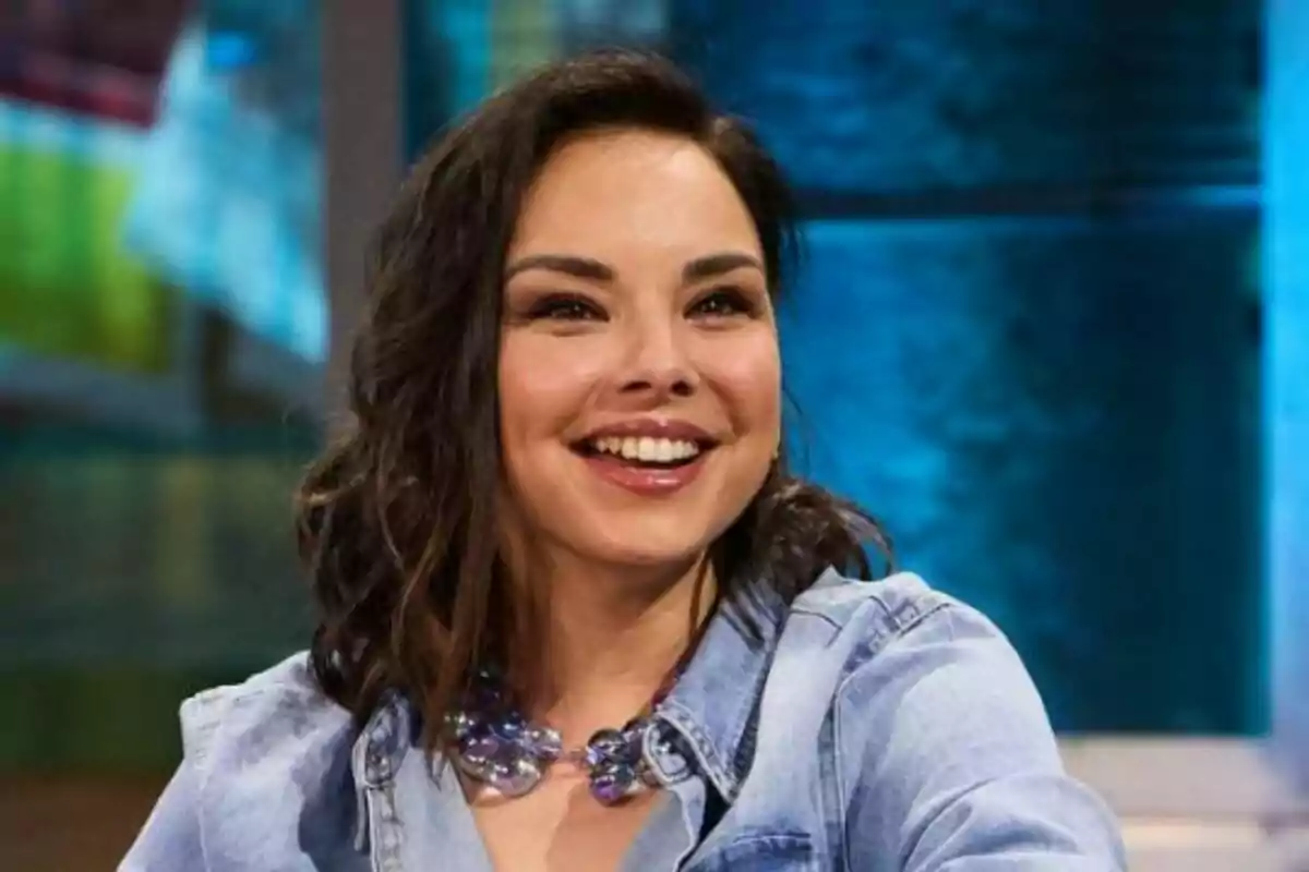 Chenoa sonriendo con una camisa de mezclilla y un collar de cuentas en un entorno interior con fondo azul en La Revuelta. Chenoa sonriendo con una camisa de mezclilla y un collar de cuentas en un entorno interior con fondo azul en La Revuelta.