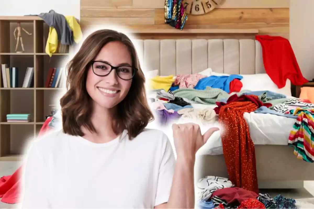 Una mujer sonriente con gafas y camiseta blanca señala con el pulgar hacia una cama desordenada llena de ropa, con un estante de libros y ropa en el fondo.