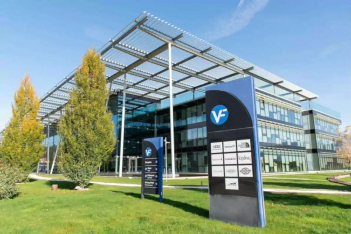 Modern building with a glass facade and exterior metal structure surrounded by grass and trees with a sign displaying various brand logos. Modern building with a glass facade and exterior metal structure surrounded by grass and trees with a sign displaying various brand logos.