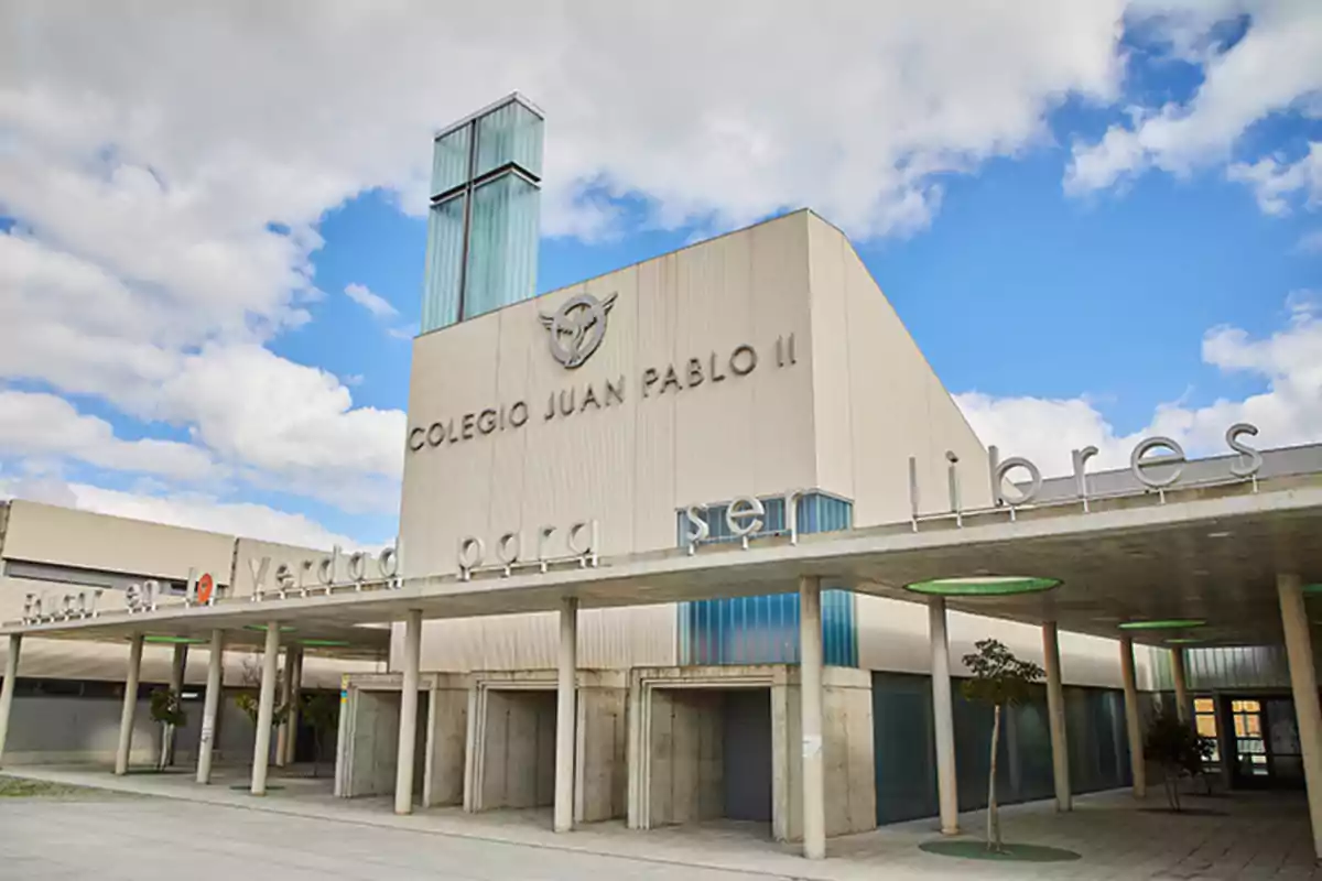 Edificio del Colegio Juan Pablo II con una cruz en la parte superior y un cielo azul con nubes de fondo.