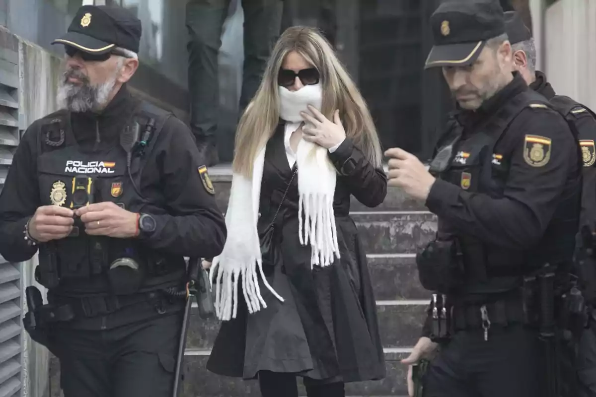 Una mujer con bufanda blanca y gafas de sol es escoltada por agentes de la Policía Nacional.