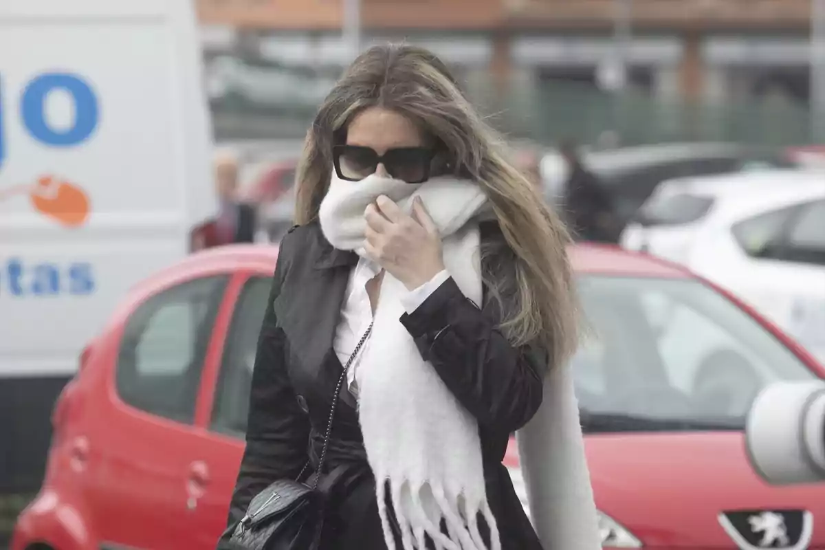 Una persona con gafas de sol y bufanda blanca caminando por la calle frente a un coche rojo. Una persona con gafas de sol y bufanda blanca caminando por la calle frente a un coche rojo.