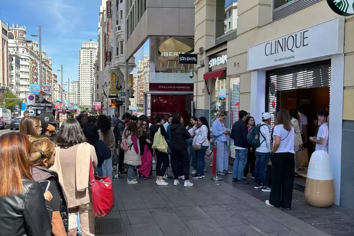 Una fila de personas espera afuera de una tienda Clinique en una calle concurrida de una ciudad, con edificios altos y varios comercios visibles en el fondo.