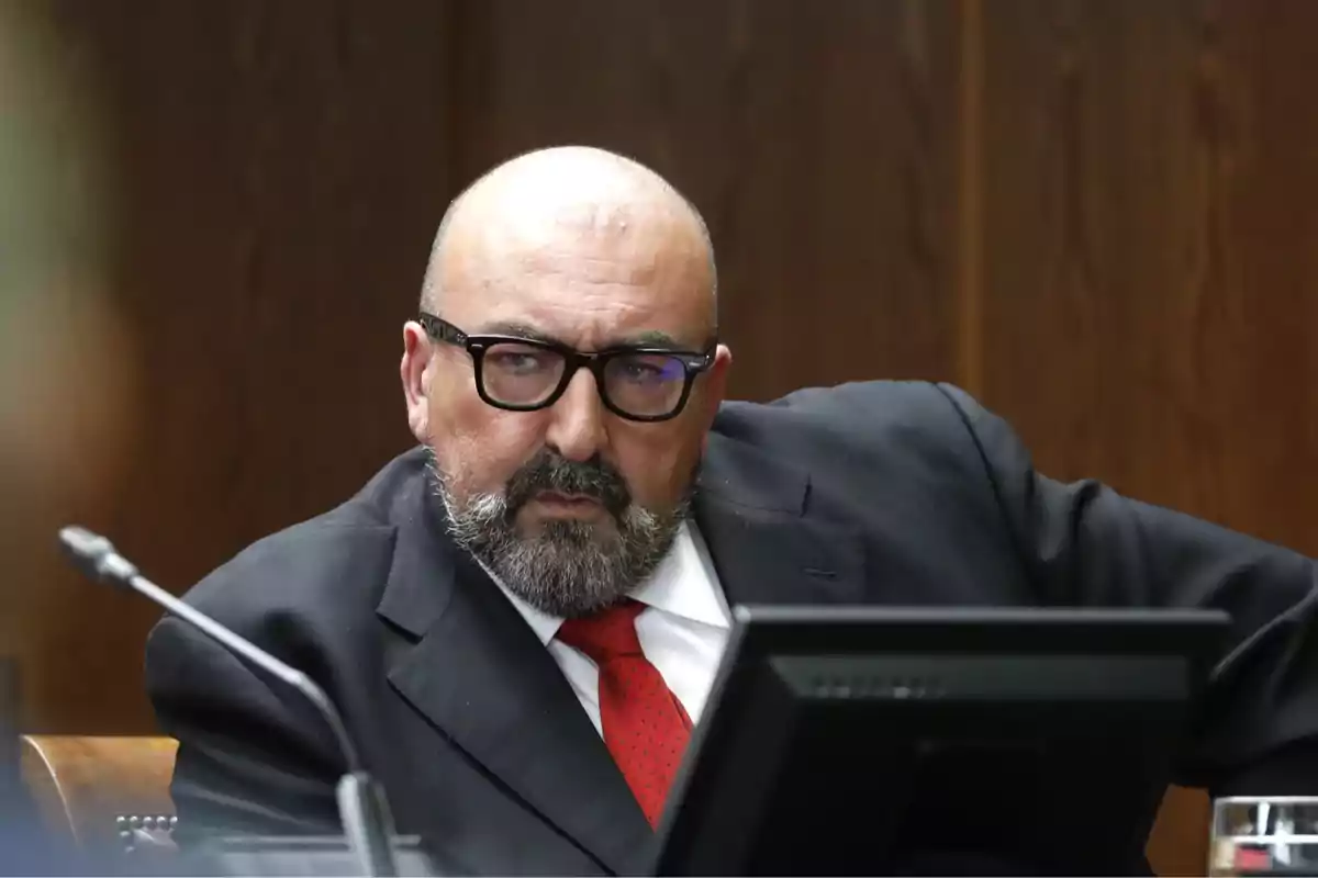 A bearded man with glasses, dressed in a dark suit and red tie, sitting in front of a screen in a formal setting. A bearded man with glasses, dressed in a dark suit and red tie, sitting in front of a screen in a formal setting.