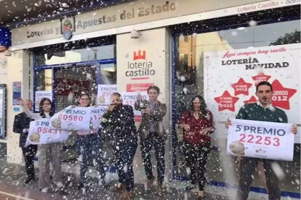 Un grupo de personas celebra frente a una administración de lotería sosteniendo carteles con números ganadores mientras cae confeti.