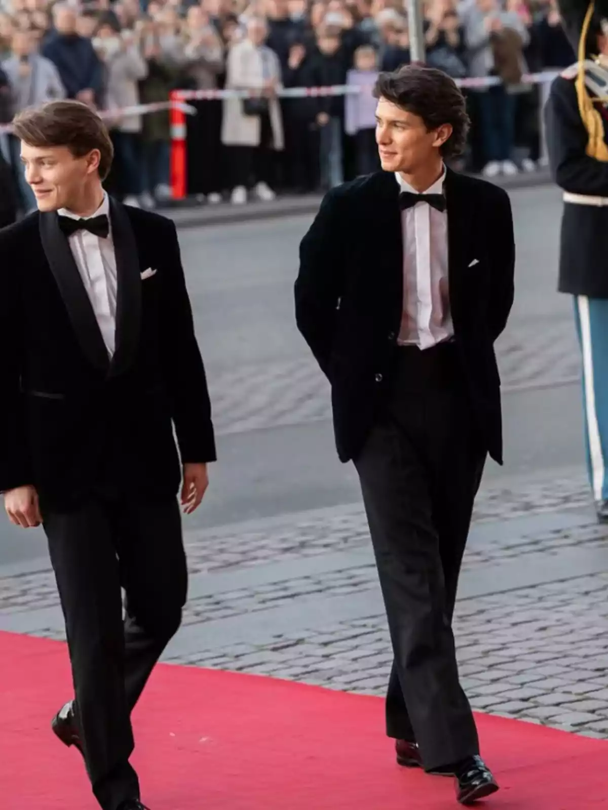 Dos hombres jóvenes con esmoquin caminan por una alfombra roja mientras una multitud observa detrás de una barrera. Dos hombres jóvenes con esmoquin caminan por una alfombra roja mientras una multitud observa detrás de una barrera.