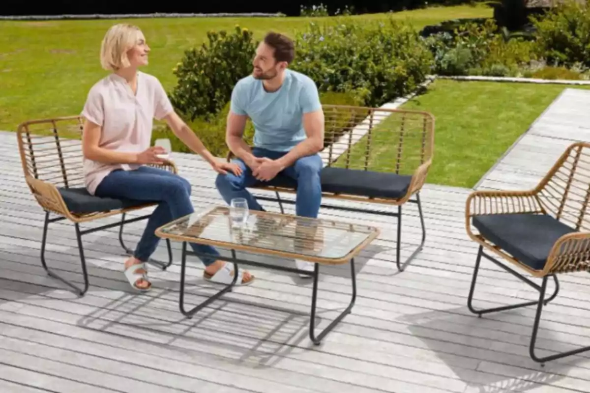 Una pareja sentada en un conjunto de muebles de exterior en una terraza de madera con césped y plantas al fondo. Una pareja sentada en un conjunto de muebles de exterior en una terraza de madera con césped y plantas al fondo.
