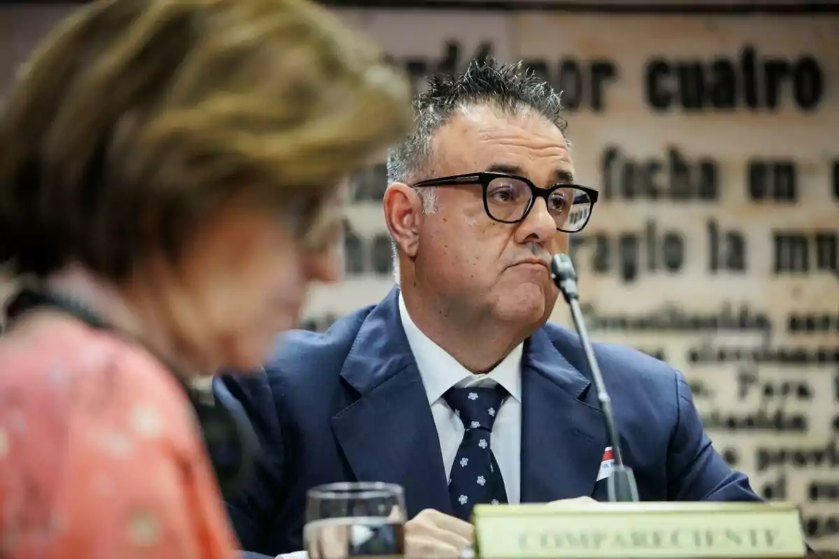 Un hombre con gafas y traje azul está sentado frente a un micrófono en una mesa, con una mujer desenfocada en primer plano.