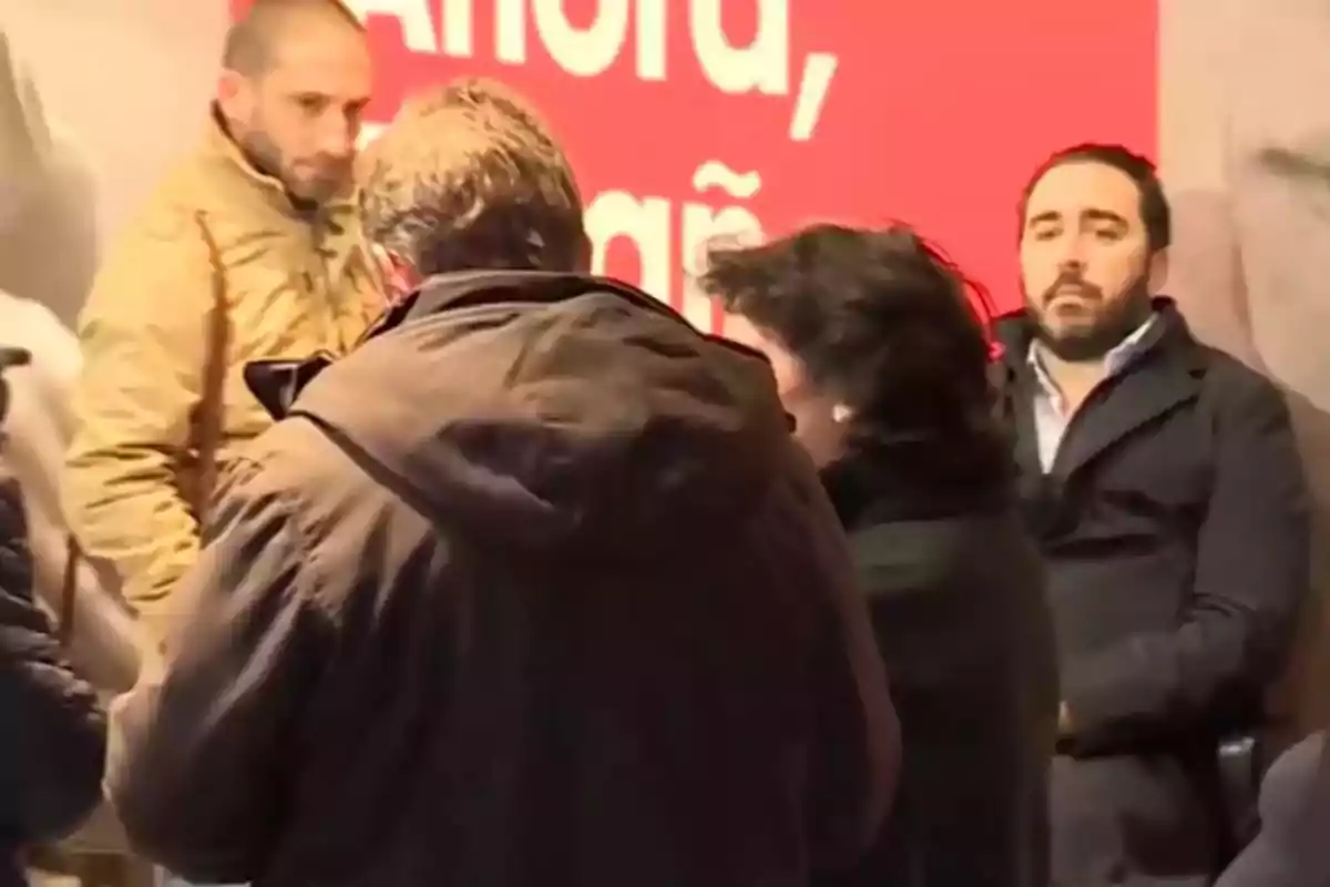 Varias personas reunidas en un espacio interior con un fondo rojo y letras blancas, algunas de ellas de espaldas y una persona sentada mirando al frente Varias personas reunidas en un espacio interior con un fondo rojo y letras blancas, algunas de ellas de espaldas y una persona sentada mirando al frente