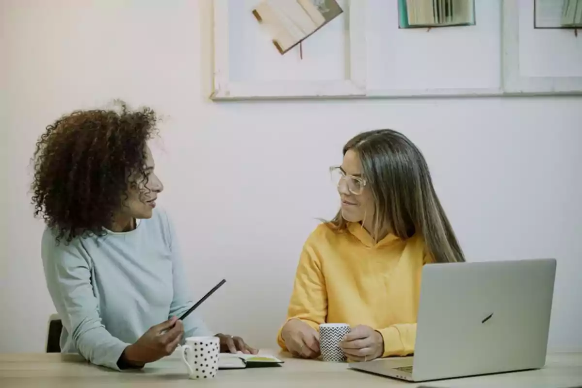Dos mujeres conversan sentadas en una mesa con tazas de café y una computadora portátil, en un ambiente relajado y profesional