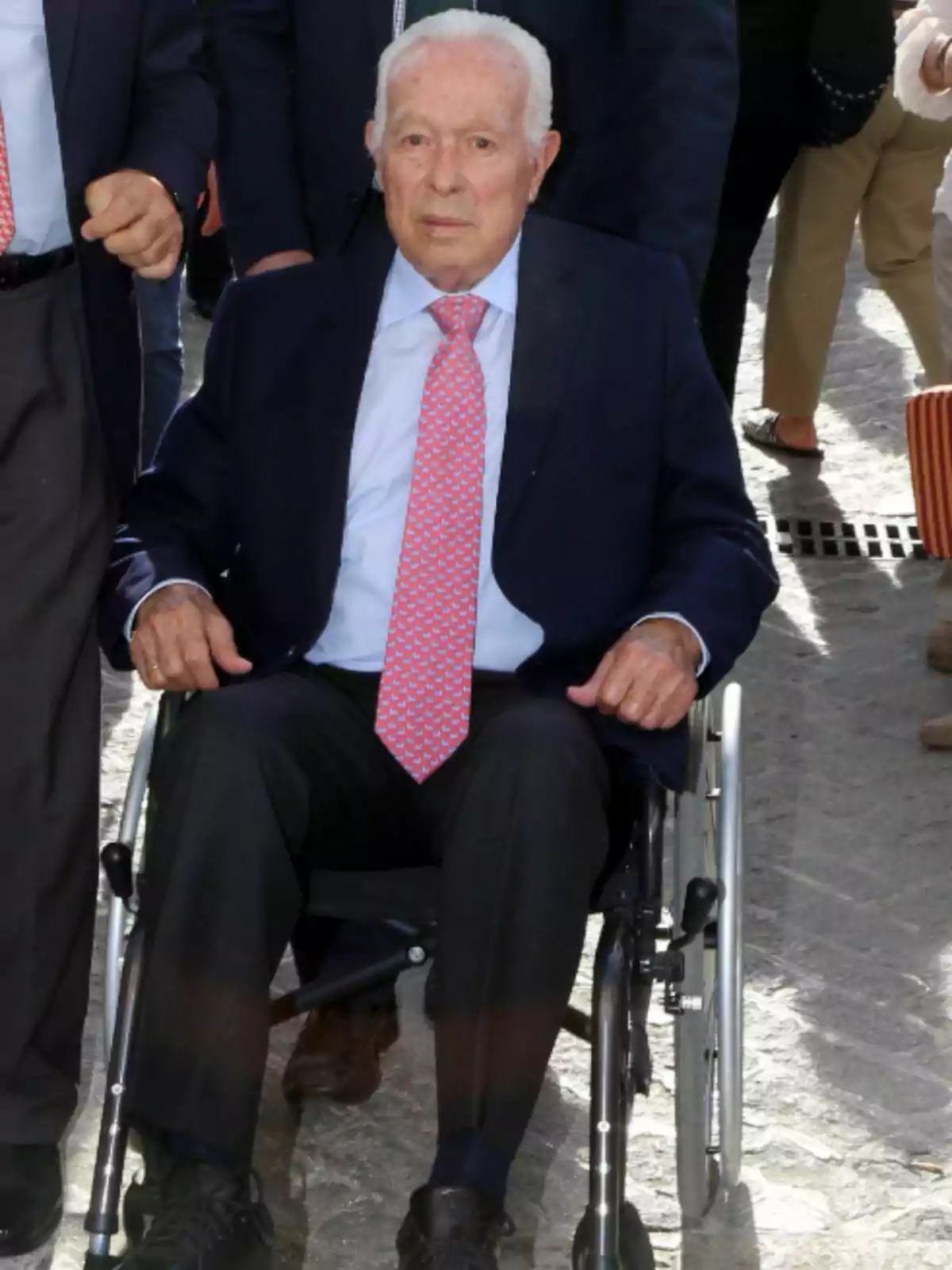 Curro Romero en silla de ruedas, vestido con traje oscuro y corbata rosa, rodeado de personas. Curro Romero en silla de ruedas, vestido con traje oscuro y corbata rosa, rodeado de personas.