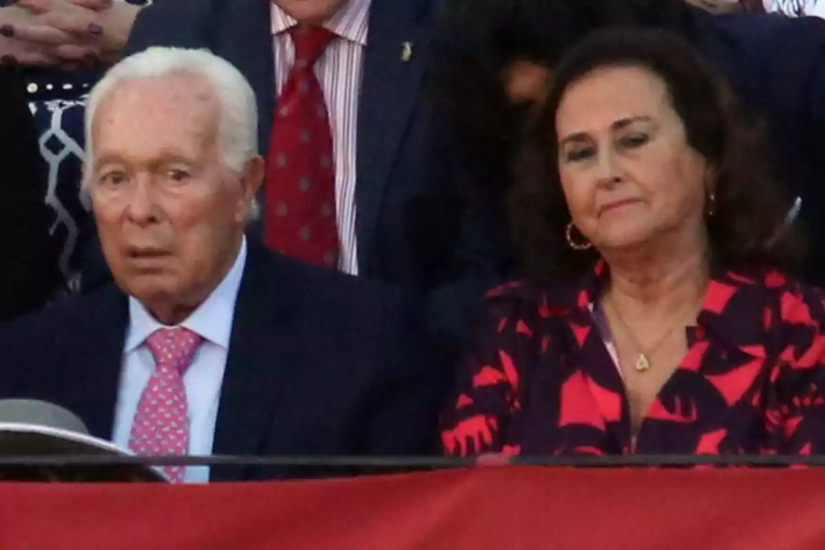 Curro Romero y Carmen Tello sentadas en un evento, una de ellas lleva traje oscuro y corbata rosa, mientras que la otra viste una blusa con estampado rojo y negro. Curro Romero y Carmen Tello sentadas en un evento, una de ellas lleva traje oscuro y corbata rosa, mientras que la otra viste una blusa con estampado rojo y negro.