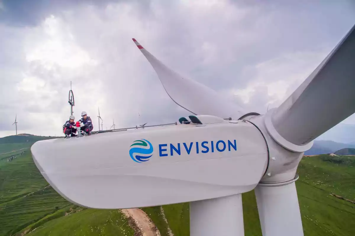 Dos personas con equipo de seguridad trabajan en la parte superior de un aerogenerador de la empresa Envision en un paisaje montañoso con más turbinas eólicas al fondo. Dos personas con equipo de seguridad trabajan en la parte superior de un aerogenerador de la empresa Envision en un paisaje montañoso con más turbinas eólicas al fondo.