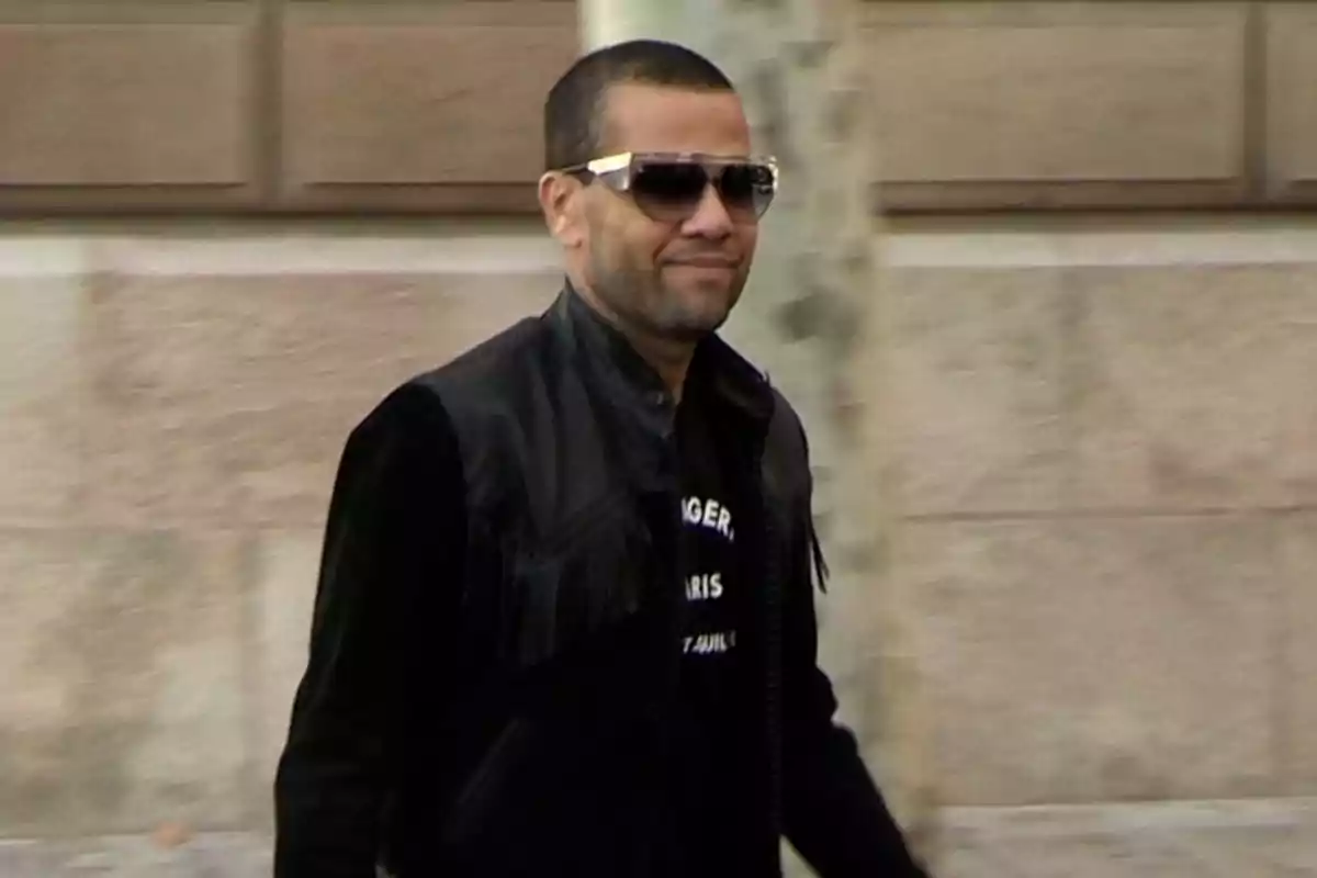 Dani Alves con gafas de sol y chaqueta negra camina al aire libre frente a una pared de piedra. Dani Alves con gafas de sol y chaqueta negra camina al aire libre frente a una pared de piedra.
