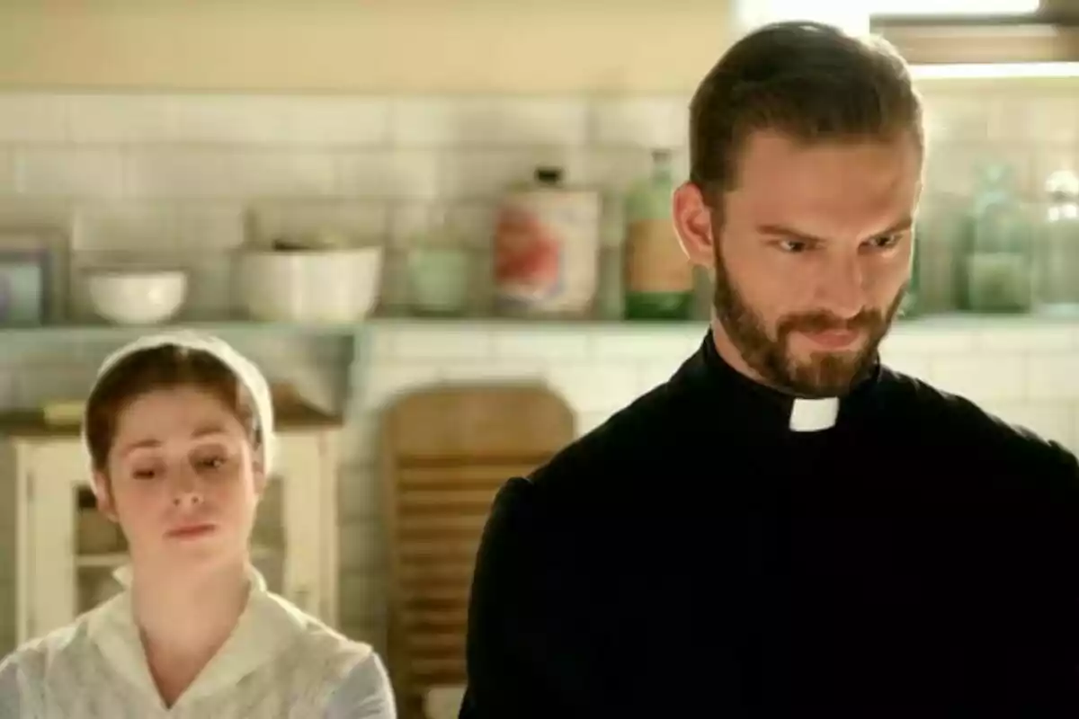 Imagen del actor Daniel Schröder vestido de sacerdote y una mujer con cofia blanca están en una cocina antigua con estantes y utensilios al fondo. Imagen del actor Daniel Schröder vestido de sacerdote y una mujer con cofia blanca están en una cocina antigua con estantes y utensilios al fondo.