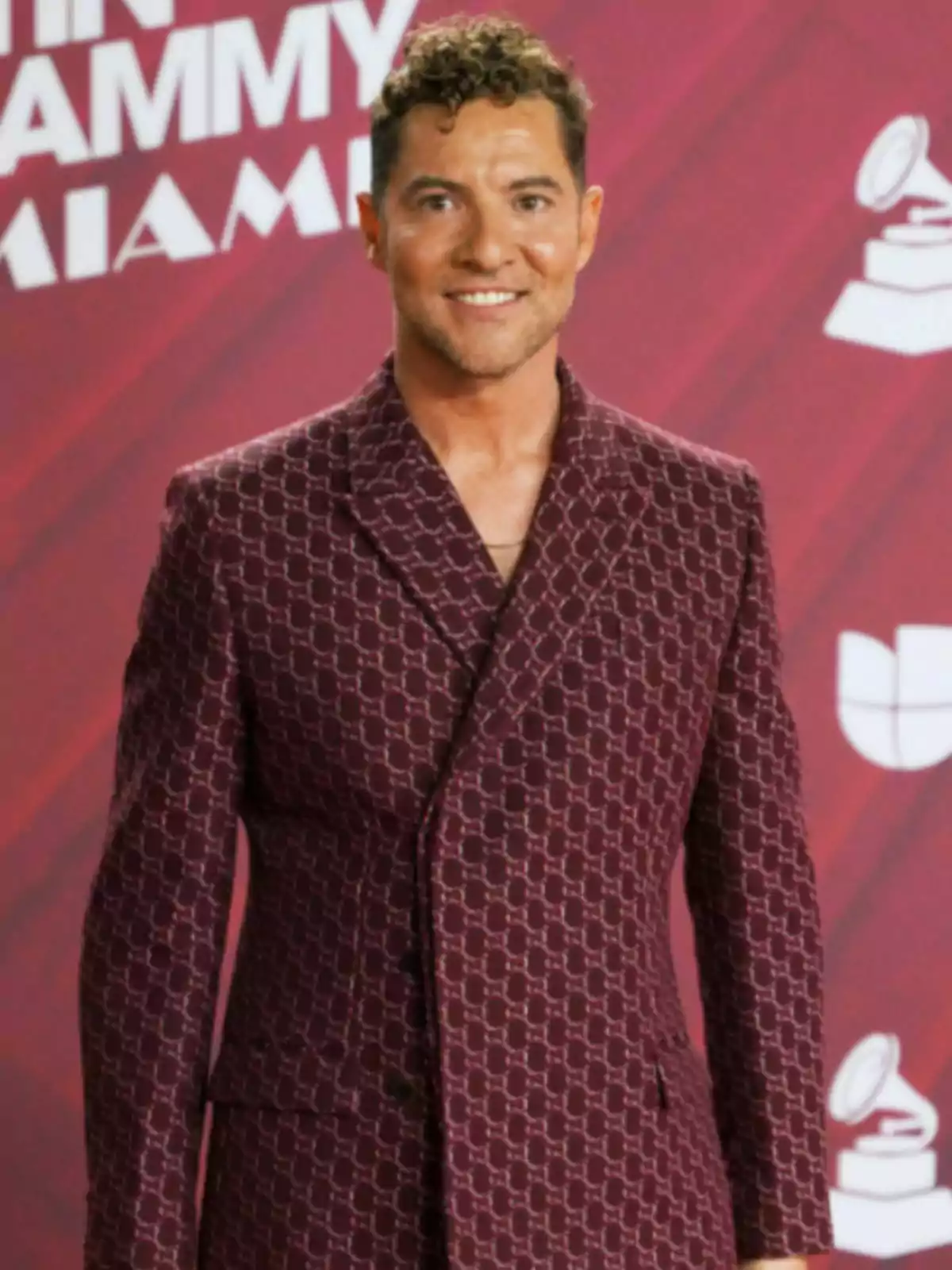 David Bisbal con un traje estampado de color burdeos posa frente a un fondo rojo con logotipos de premios. David Bisbal con un traje estampado de color burdeos posa frente a un fondo rojo con logotipos de premios.
