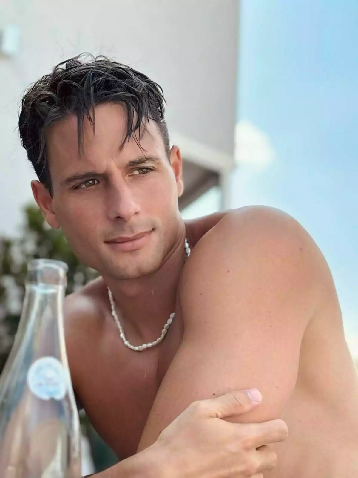 David Matarín sin camisa con collar de perlas, sentado al aire libre junto a una botella de vidrio. Post en Instagram