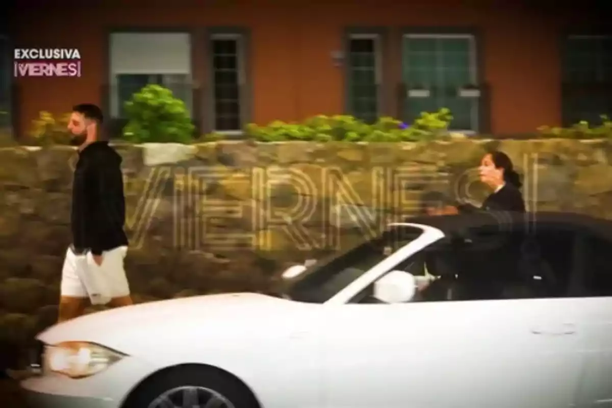 David Rodríguez walks on the sidewalk next to a white convertible car, while Anabel Pantoja follows some distance behind him.