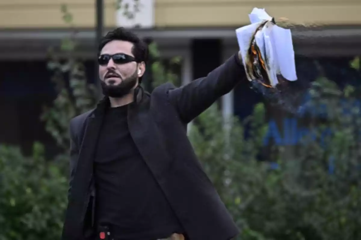 Un hombre con gafas de sol sostiene papeles en llamas al aire en un entorno al aire libre.