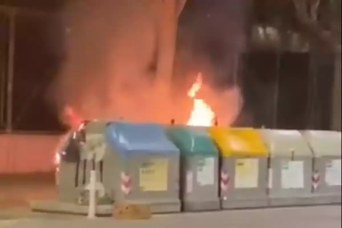 Contenedores de reciclaje en la calle con un incendio en el fondo.