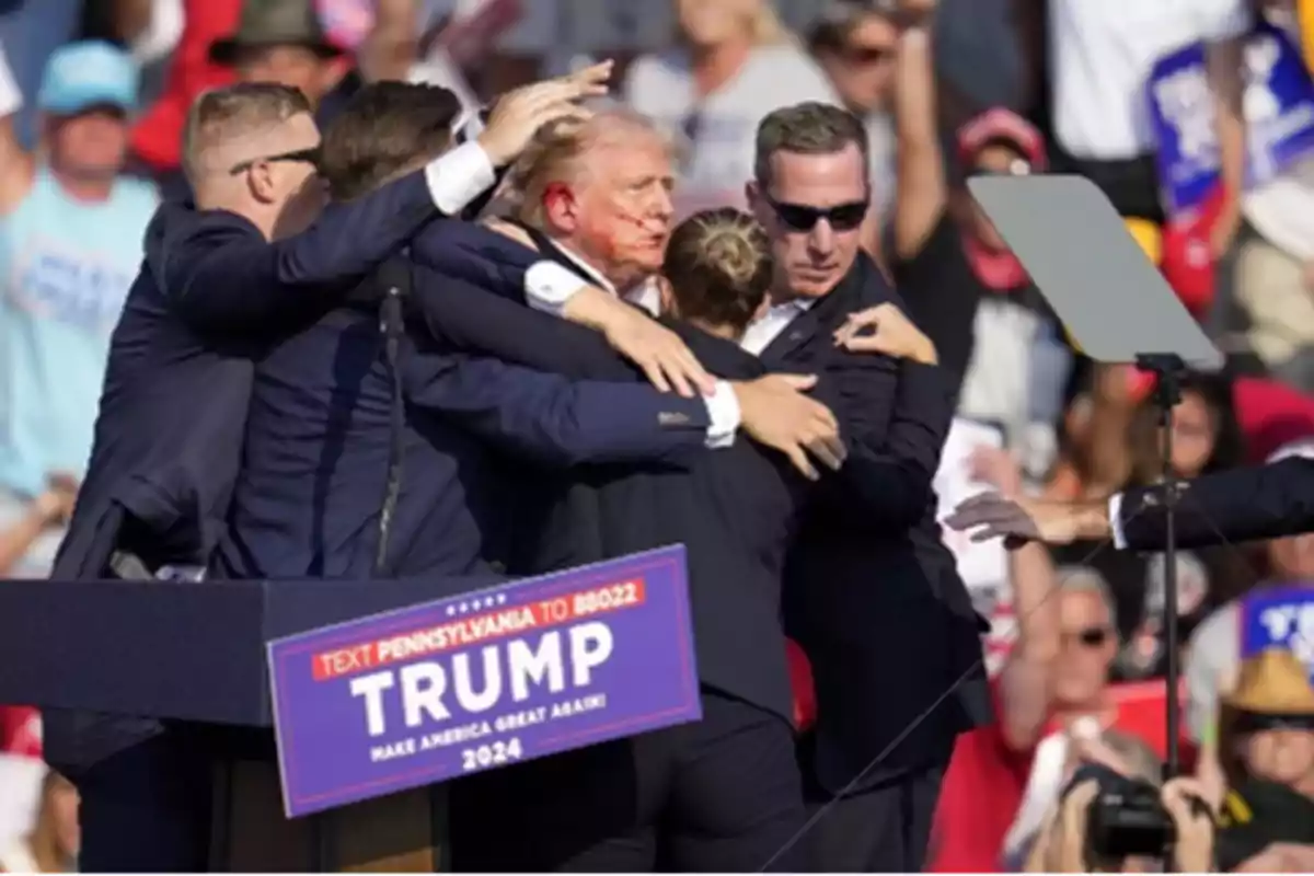 Un grupo de personas rodea y protege a un hombre en un mitin político con un cartel que dice "TEXT PENNSYLVANIA TO 88022 TRUMP MAKE AMERICA GREAT AGAIN! 2024".