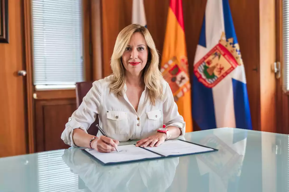 Mujer rubia firmando un documento en una oficina con banderas de fondo.