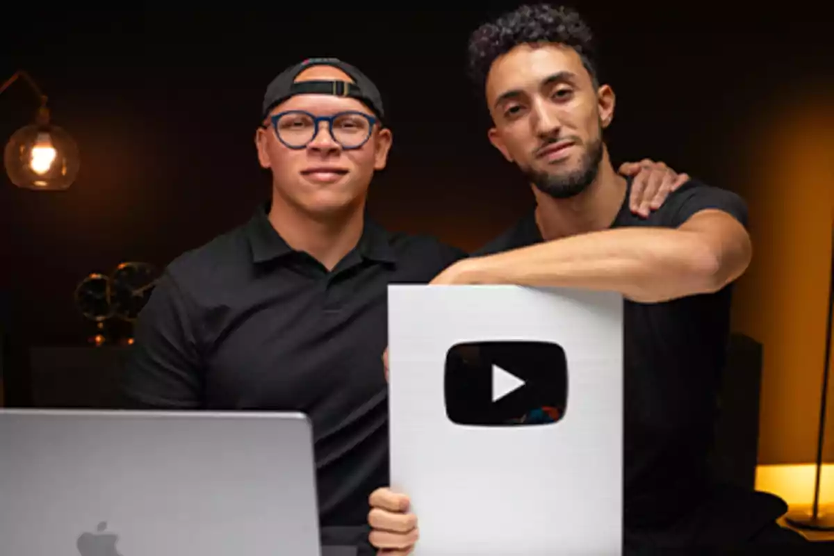 Two people posing with a YouTube silver play button and a laptop on a table.