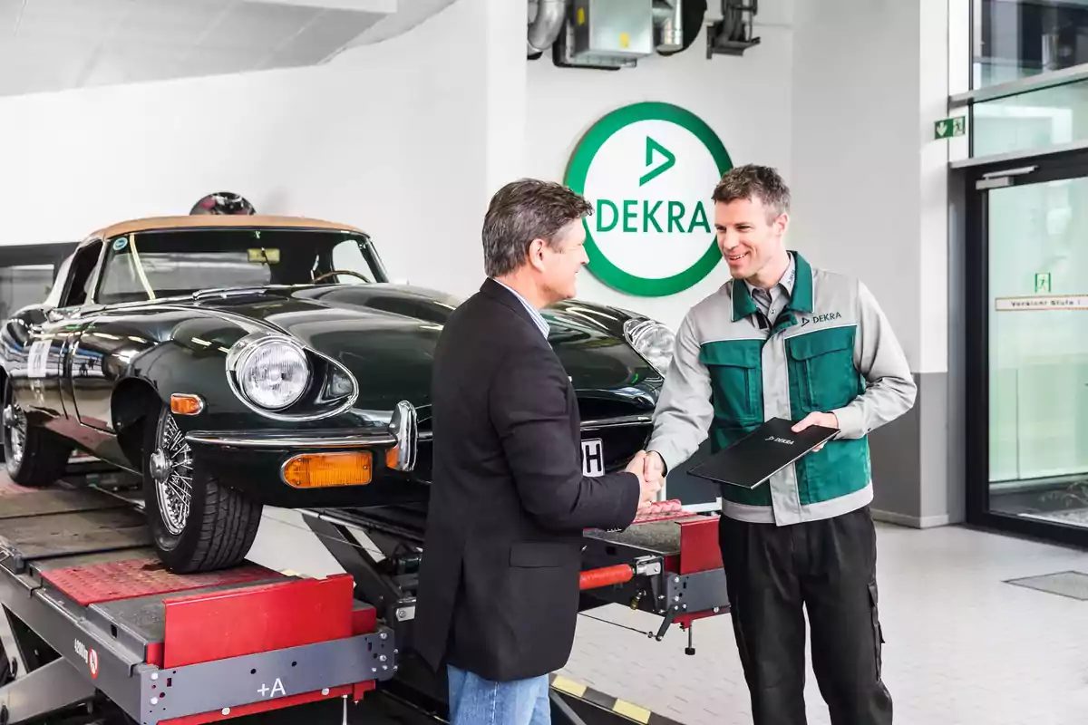 Dos hombres estrechándose la mano en un taller de automóviles con un coche clásico en una plataforma elevadora y un logotipo de DEKRA en la pared.