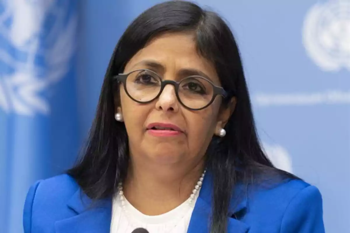 Mujer con gafas y cabello oscuro hablando frente a un micrófono con un fondo azul.